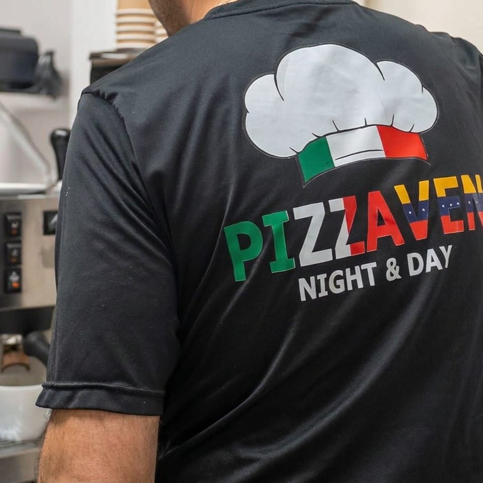 Restaurant worker wearing a black Pizza Venti Night & Day shirt works beside a coffee machine in a tiled kitchen.