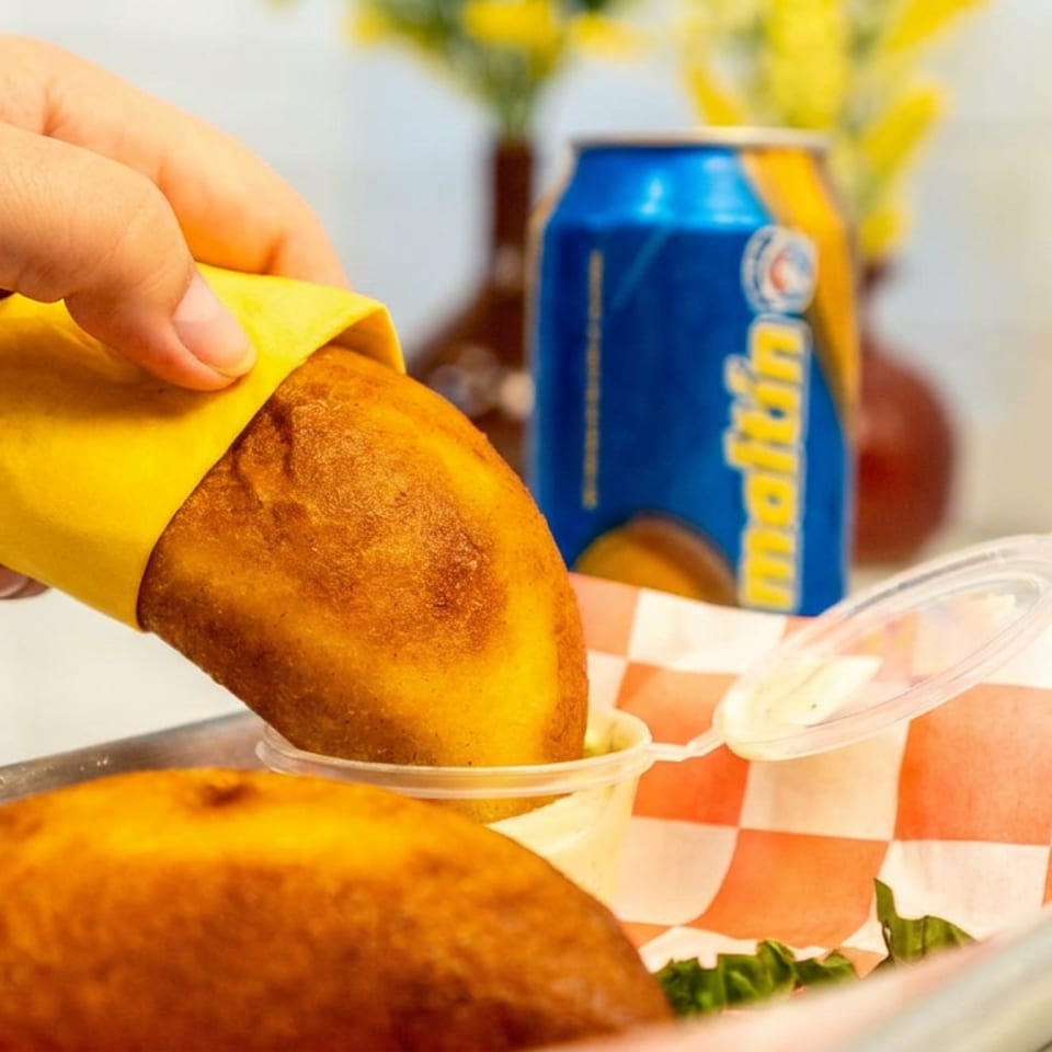 Hand dips a golden fried pastry into sauce above a checkered-lined tray, with another pastry and soda can nearby.