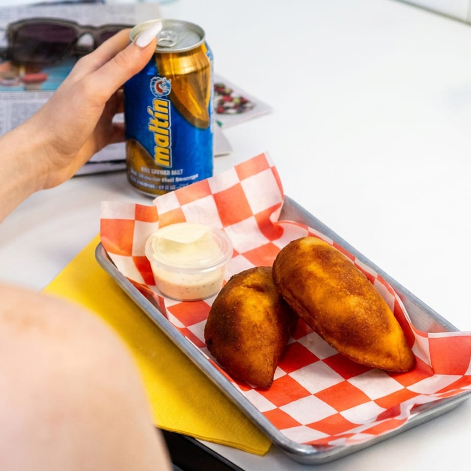 Tray of two golden fried empanadas with dipping sauce and a Matin beer can on a restaurant table.