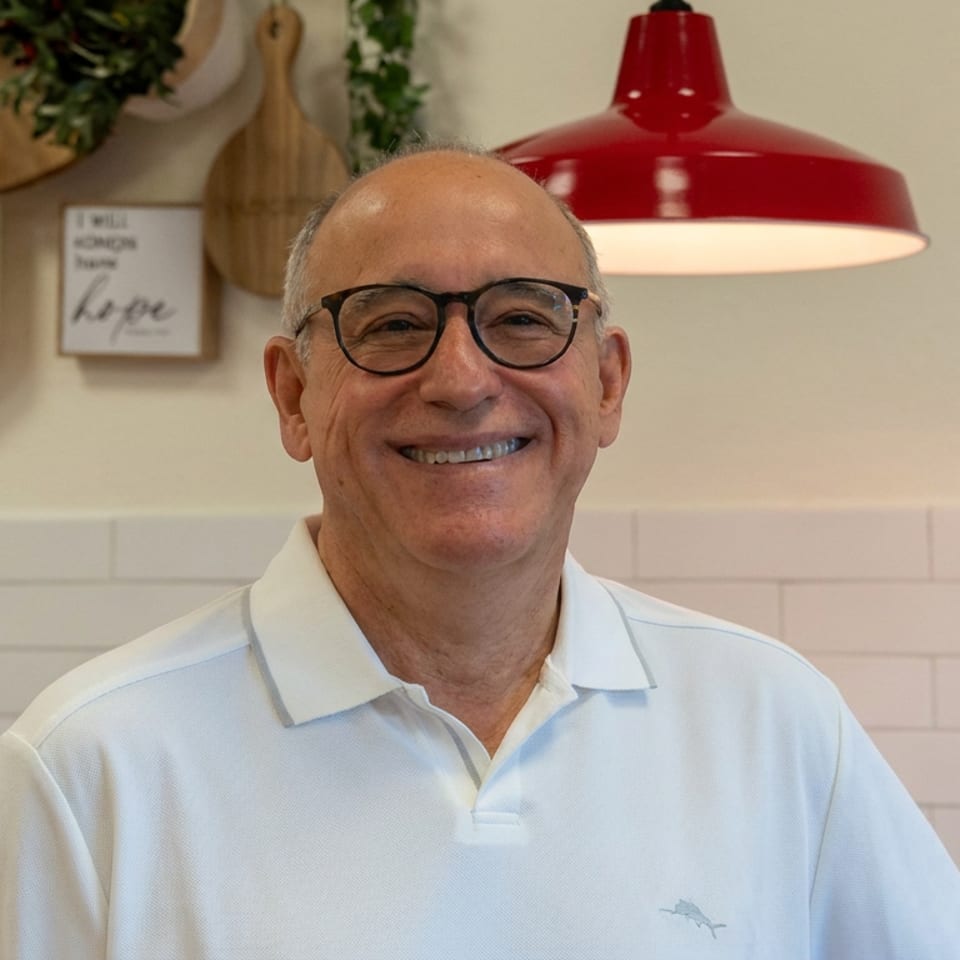 Smiling man wearing glasses and a white polo shirt stands in a warmly lit pizzeria beneath a red pendant lamp.