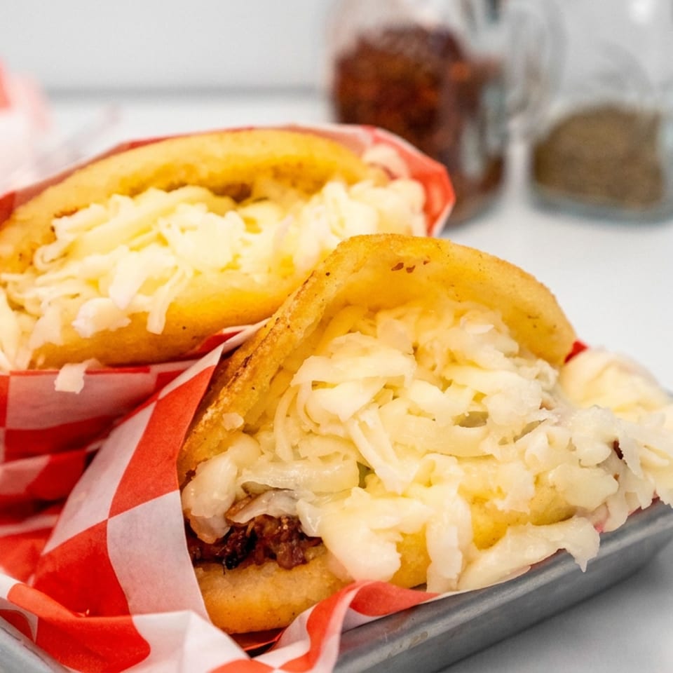 Two golden stuffed corn cakes filled with melted shredded cheese and meat, served on red-and-white checkered paper in a tray.