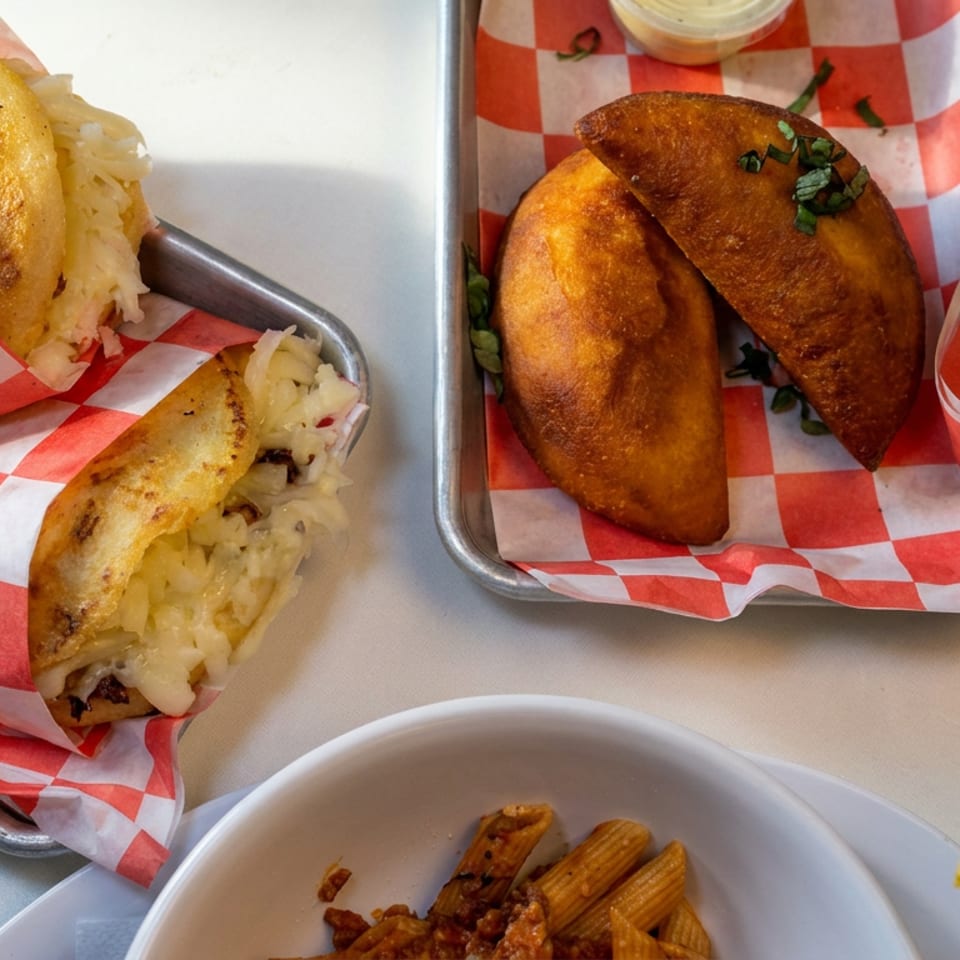 Two trays display golden stuffed pastries and empanadas on red-and-white checkered paper, with churros in a bowl nearby.