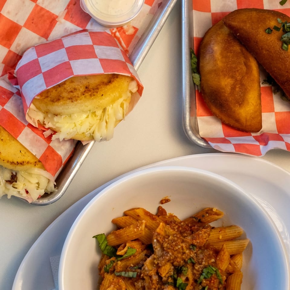 Overhead spread of stuffed golden pastries, filled corn cakes, and penne pasta with meat sauce on checkered-lined trays.