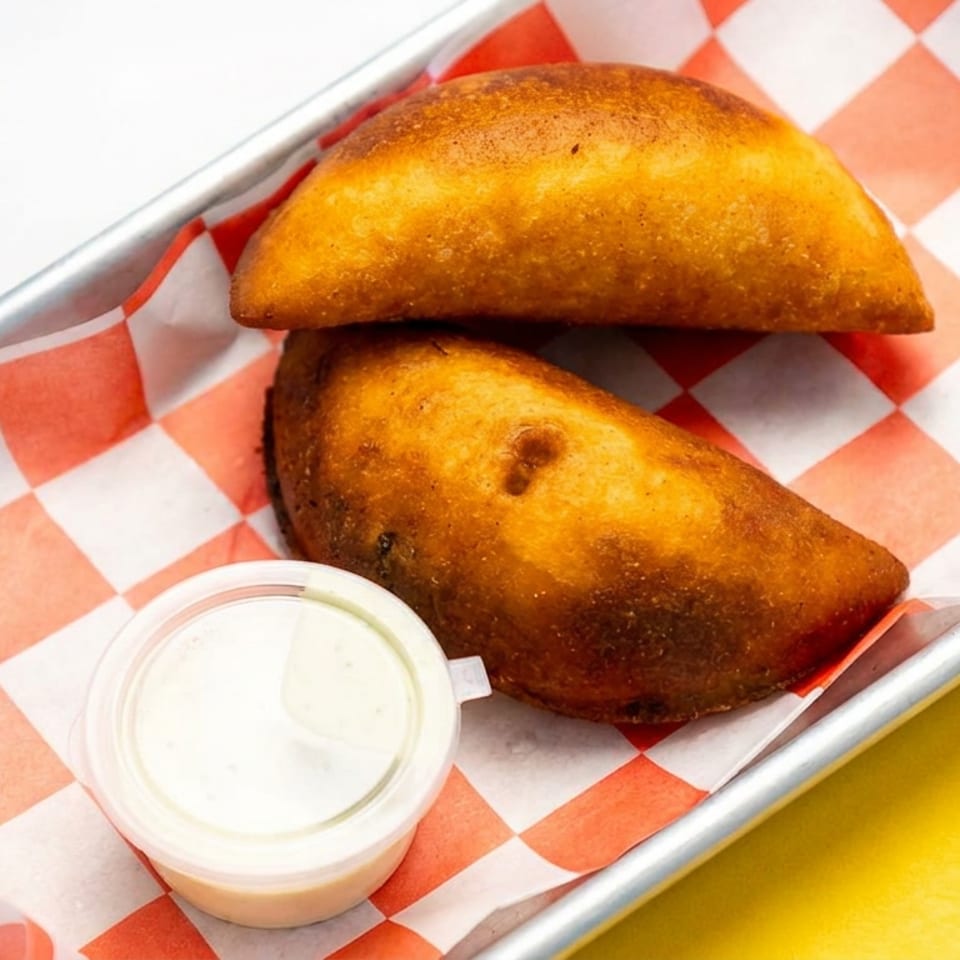 Two golden-brown empanadas served with creamy dipping sauce on red-and-white checkered paper in a metal tray.
