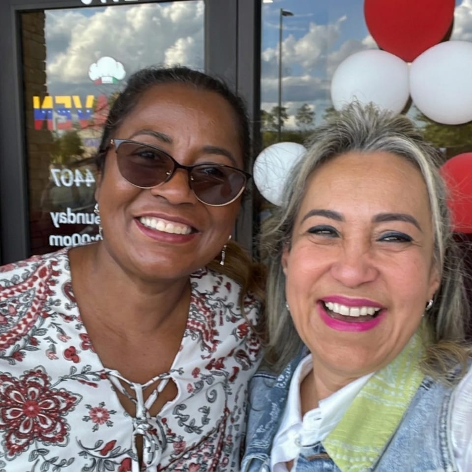 Two smiling women pose outside a restaurant entrance decorated with red and white balloons.